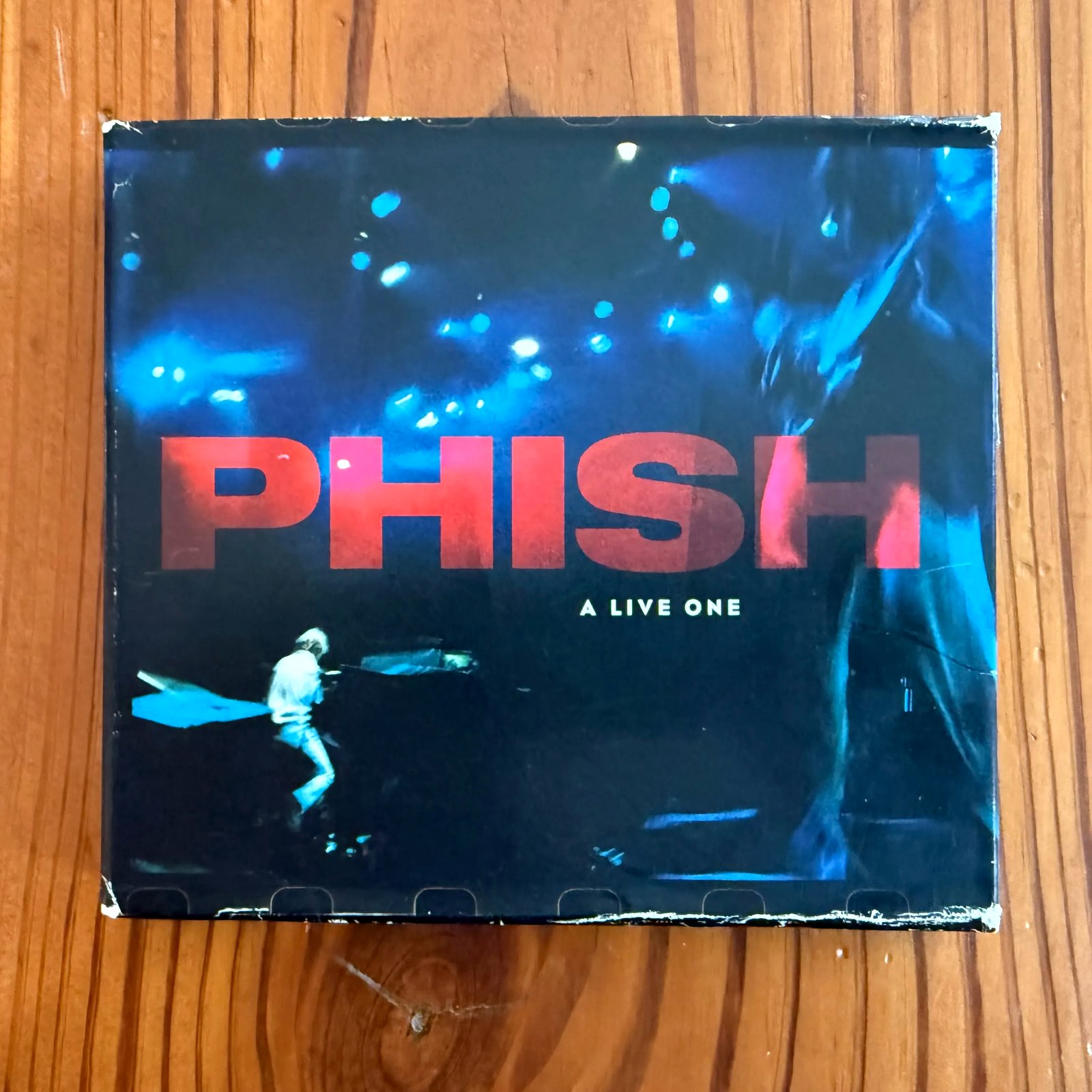 A worn Phish CD, "A Live One," lying face up on a wooden tabletop. The cover is a dark blue concert photograph with the band's name across the middle in large red capitals and the album title below it in small white letters. A figure in white, Page McConnell, is bent over a piano at the lower left, lit by blue stage lights. The cardboard slipcase is frayed and split at all four corners.